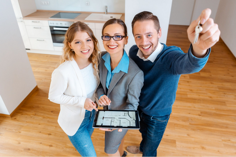 Three people stand in an empty room with one holding a key and another holding a tablet displaying a floor plan.