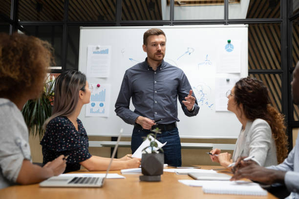 Businessman presenting investment strategies to a team in a conference room with charts and graphs on whiteboard.