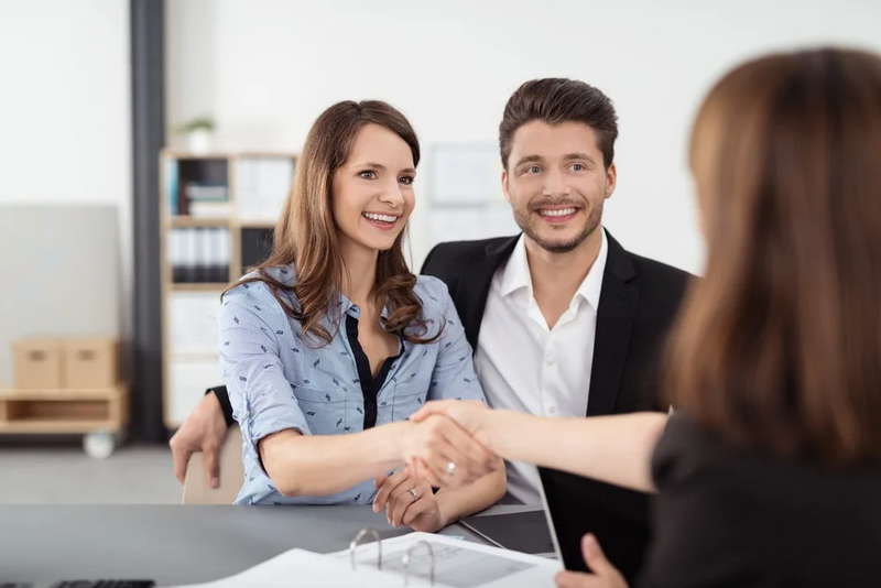 A smiling couple shakes hands with a person across a desk in an office setting.