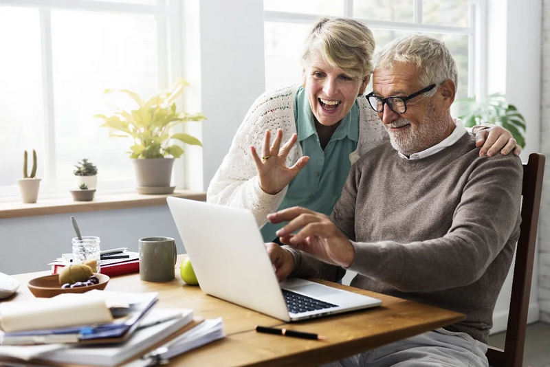 An elderly couple smiles and waves at a laptop screen during a video call in a cozy, well-lit room.