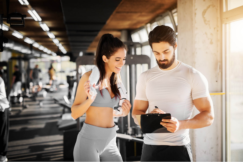 A woman and a man are conversing while standing in a gym, with the man holding a clipboard.