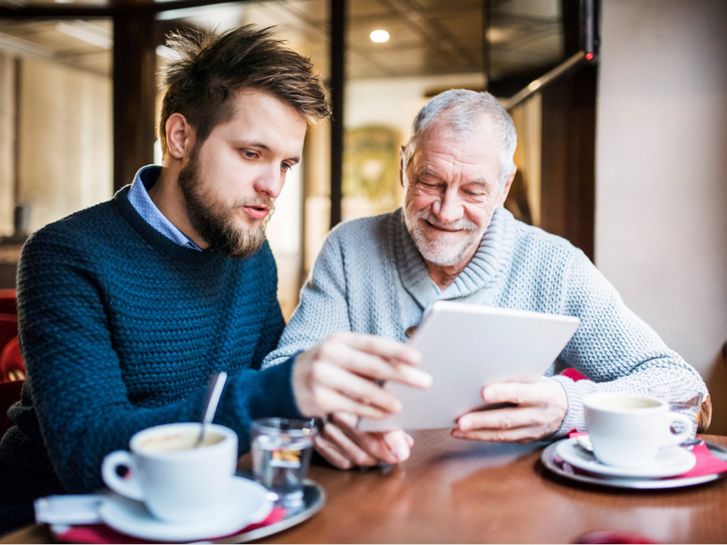 A young man and an elderly man are looking at a tablet together in a café.