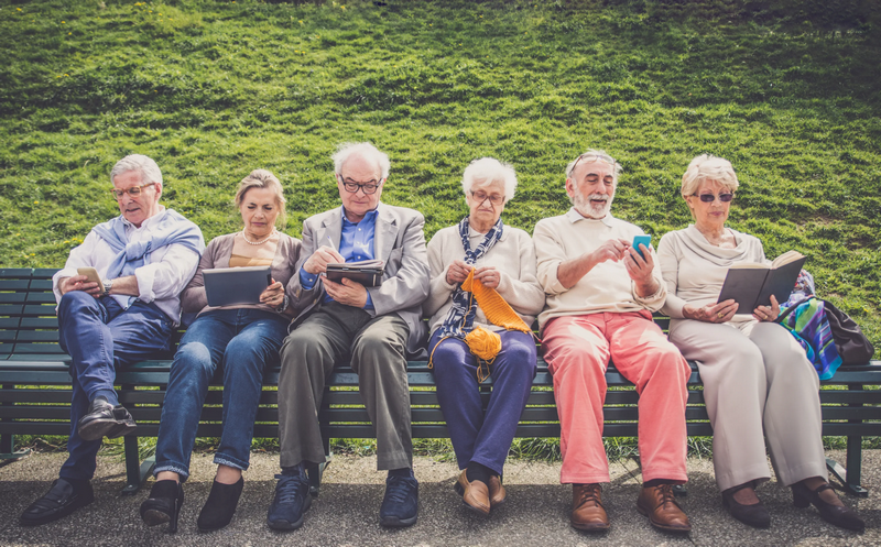 A group of elderly people sitting on a bench, engaging with various activities such as reading, knitting, and using electronic devices.